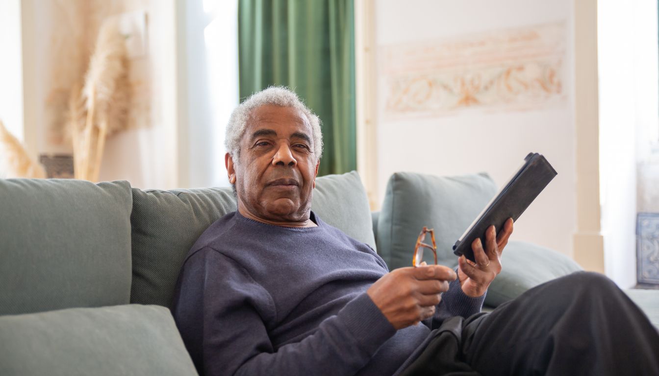 An older man sitting on his sofa holding a tablet and reading glasses